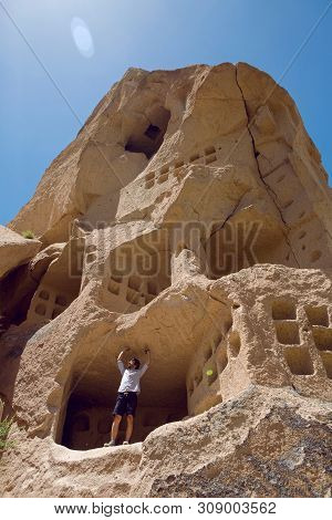 Guy In Shirt And Shorts Stand On A Rock House With Windows In Uchhisar