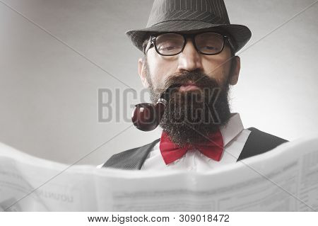 The Elegant Old-fashioned Bearded Hipster Middle Aged Man With Tobacco Pipe Is Reading Newspaper.