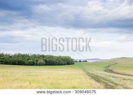 Summer Landscape In Cloudy Day. Dirty Road Cross The Green Meadow With Wild Herbs. Mountain On A Lak
