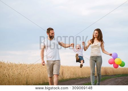 Happy Family Walking In The Field. Mom, Dad And Son Walk Outdoors, Parents Holding The Baby Boys Han