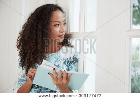 Portrait Of Cheerful Young Asian Woman With Book Of Poen Looking Through Window Of Her Bedroom