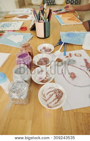 Jars Of Colorful Glitter, Plastic Plater And Paper Sheets With Drawings On Wooden Table In Art Schoo