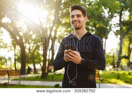 Image of a cheerful optimistic young sports fitness man standing in green park nature using mobile phone chatting listening music.