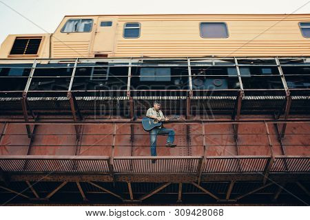 Bluesman With Acoustic Guitar On Railroad Bridge. Cool Guy Plays Guitar Near Running Train. Showy Pe