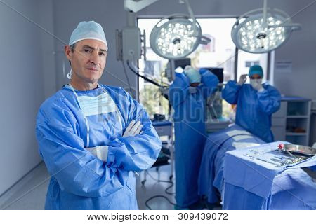 Portrait of Caucasian male surgeon standing with arm crossed while diverse surgeons performing surgery in operation room while surgeons getting ready for operation at hospital