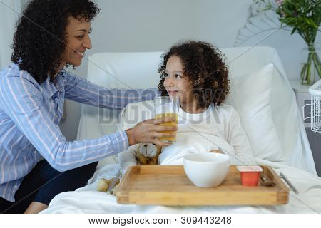 Side view of happy mixed race mother feeding her son orange juice in bed in the ward at hospital