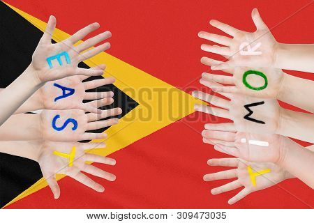 Inscription East Timor On The Children's Hands Against The Background Of A Waving Flag Of The East T
