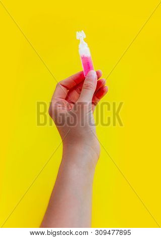 Female Hand Holding One Pink Liquid Medicine In An Ampoule, Isolated On Yellow Background.