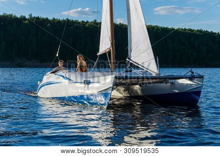 Couple Rest On A Small Sailboat Catamaran Going Against The Wooded Shore