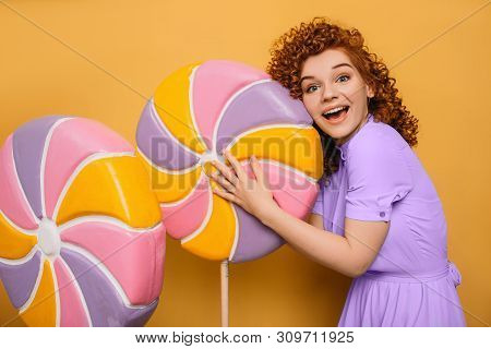 Mmm Yummy It Is Very Sweet Lollipop. Curly Red-haired Woman Hugging Candy On Yellow Background
