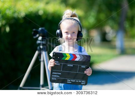 A Little Girl In Headphones With A Tv Numerator In Her Hands Is Standing Next To The Camera On A Tri