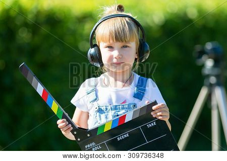 Little Girl In Headphones With A Tv Numerator In Her Hands Is Standing Next To The Camera On A Tripo