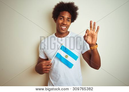 Afro american tourist man holding Argentinian Argentina flag over isolated white background doing ok sign with fingers, excellent symbol