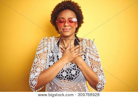 Young african american woman with afro hair wearing bikini and heart shaped sunglasses smiling with hands on chest with closed eyes and grateful gesture on face. Health concept.