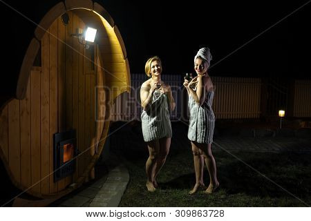 Two Girlfriends Women Drinking Red Wine From Glasses Outsite Near A Wooden Mobile Sauna Wearing Just