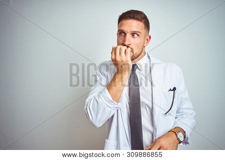 Young handsome doctor man wearing white profressional coat over isolated background looking stressed and nervous with hands on mouth biting nails. Anxiety problem.