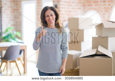 Middle age woman moving to a new house arround cardboard boxes doing happy thumbs up gesture with hand. Approving expression looking at the camera with showing success.