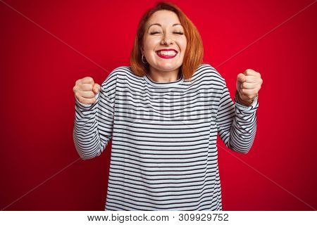 Young redhead woman wearing strapes navy shirt standing over red isolated background excited for success with arms raised and eyes closed celebrating victory smiling. Winner concept.