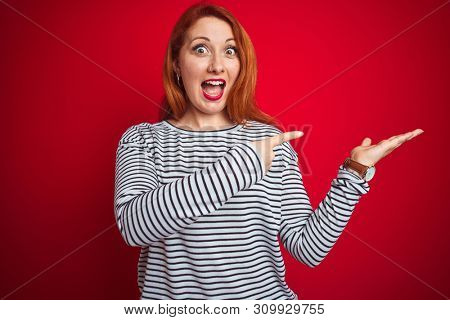 Young redhead woman wearing strapes navy shirt standing over red isolated background amazed and smiling to the camera while presenting with hand and pointing with finger.