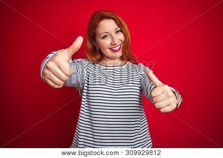 Young redhead woman wearing strapes navy shirt standing over red isolated background approving doing positive gesture with hand, thumbs up smiling and happy for success. Winner gesture.