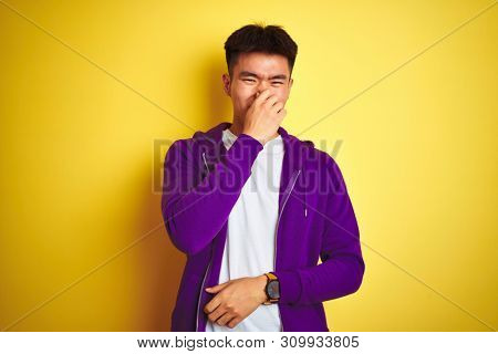 Young asian Chinese man wearing purple sweatshirt standing over isolated yellow background smelling something stinky and disgusting, intolerable smell, holding breath with fingers on nose. Bad smells