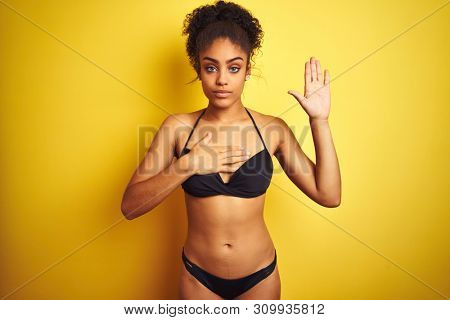 African american woman on vacation wearing bikini standing over isolated yellow background Swearing with hand on chest and open palm, making a loyalty promise oath