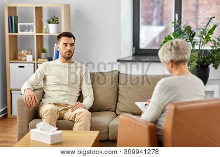 psychology, mental therapy and people concept - senior woman psychologist taking notes to notebook and sad young man patient at psychotherapy session