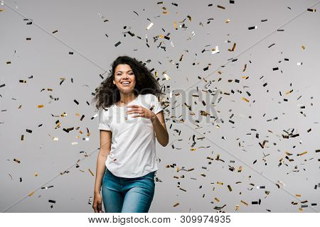 Young African American Woman Smiling Near Shiny Confetti While Standing On Grey