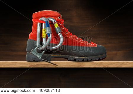 Mountaineering Equipment On A Wooden Shelf. An Old Hiking Boot, Two Carabiners And A Climbing Piton 