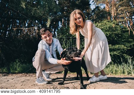 Lovely Couple Is Walking With A Black Labrodor On A Leash. Lovers Smile And Watch The Dog. A Walk In
