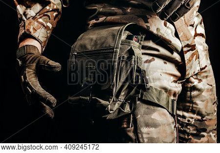 Photo Of Soldier In Camouflaged Uniform And Tactical Gloves Using Leg Bag On Black Background.