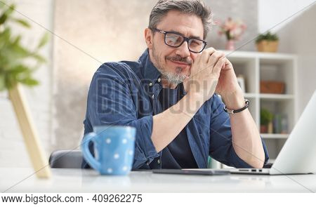 Bearded man working online with laptop computer at home sitting at desk. Home office, browsing internet, study room. Portrait of mature age, middle age, mid adult man in 50s.