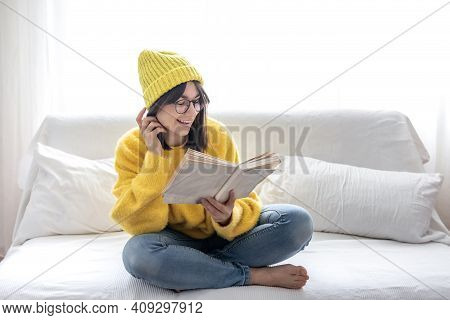 Stylish Beautiful Brunette Girl In Glasses And A Hat Reads A Book On The Sofa. Self-development And 