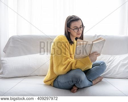 Stylish Brunette Girl In A Yellow Sweater And Glasses Reads A Book At Home On The Couch. The Concept