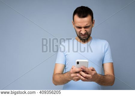 Photo Of Dissatisfied Handsome Young Man With Beard Wearing Everyday Blue T-shirt Isolated Over Blue