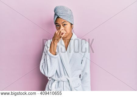 Young chinese woman wearing shower towel cap and bathrobe smelling something stinky and disgusting, intolerable smell, holding breath with fingers on nose. bad smell 