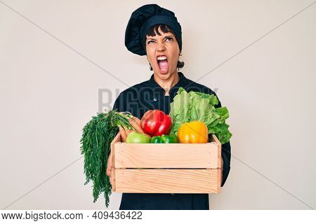 Beautiful brunettte woman chef holding fresh veggetables angry and mad screaming frustrated and furious, shouting with anger. rage and aggressive concept. 