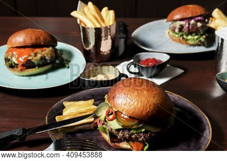 Different Burgers On The Table, Served Frenc Fries And Sauces On Plates. The Closest Spicy Beef Burg