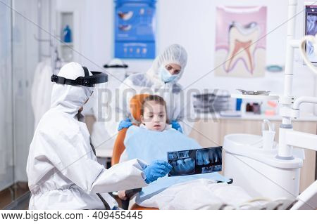 Dentist Dressed In Ppe Suit Reviewing Panoramic Mouth Radiography Of Little Girl Patient. Stomatolog