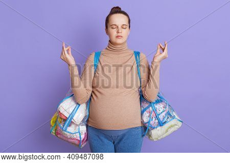 Calming Pregnant Woman Isolated Over Lilac Background, Keeps Eyes Closed, Meditates, Trying To Relax