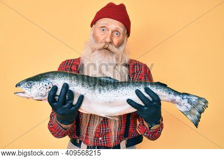 Old senior man with grey hair and long beard showing raw salmon in shock face, looking skeptical and sarcastic, surprised with open mouth 