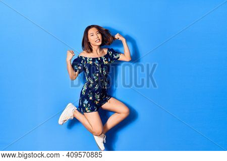 Young beautiful chinese woman wearing casual clothes smiling happy. Jumping with smile on face doing strong geture over isolated blue background