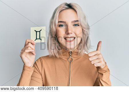 Young blonde girl holding paper with aquarius zodiac sign smiling happy and positive, thumb up doing excellent and approval sign 