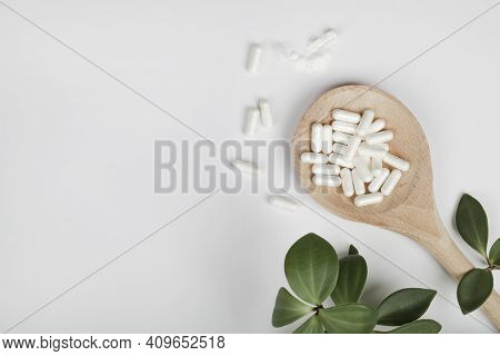 Herbal Pills In Wooden Spoon And Green Plant Leaves On White Background. Alternative Medicine And He