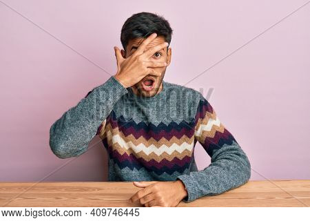 Young handsome man wearing casual winter sweater sitting on the table peeking in shock covering face and eyes with hand, looking through fingers afraid 