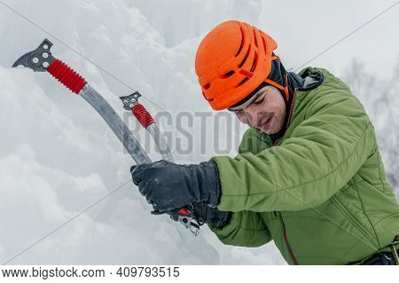 Athletic Alpinist Man In Orange Helmet And Ice Tools Axe Climbing A Large Wall Of Ice. Outdoor Sport