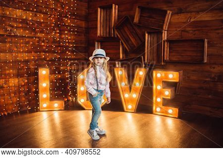 Portrait Of A Little Girl Wearing A Hat On A Background Of Wooden Letters Decorated With Lamps
