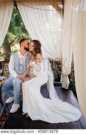 Groom In Blue Suit And Bride With Long Curls With Bouquet Sit In A Gazebo
