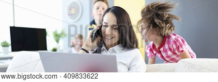 Woman Is Sitting On Couch Holding Laptop And Talking On Smartphone Next To The Children Are Playing.