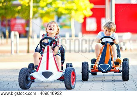 Two Active Little Kid Boys Driving Pedal Race Car In Summer Garden, Outdoors. Children, Best Friends
